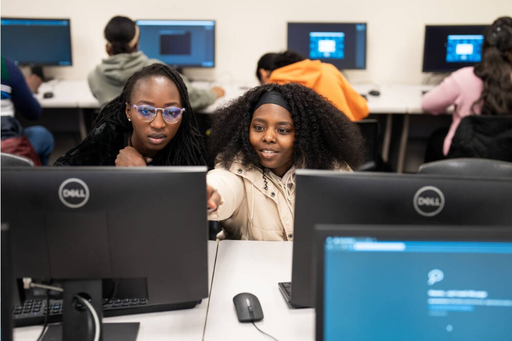 Two females look at the monitor of a computer with one pointing to it. Other computers are visible within the room that looks like a computer lab.